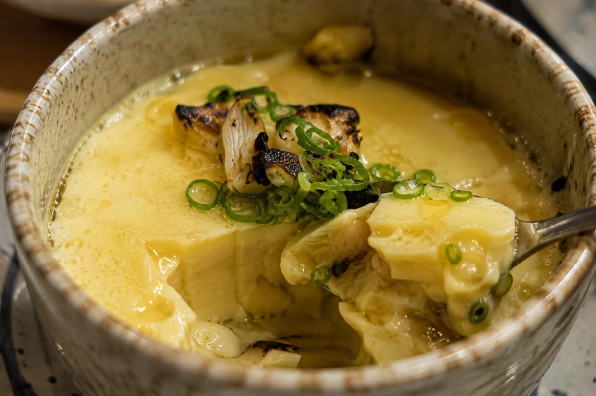 An extreme macro close-up shot reveals the interior texture of the steamed chawanmushi as a metal spoon scoops out a soft, silky portion of the pale yellow egg custard. The top layer displays a glossy sheen from droplets of ginger oil, topped with charred leek chunks and vibrant green scallion rings. The break in the custard highlights its delicate, smooth consistency and the hidden savory filling underneath. An extreme macro close-up shot reveals the interior texture of the steamed chawanmushi as a metal spoon scoops out a soft, silky portion of the pale yellow egg custard. The top layer displays a glossy sheen from droplets of ginger oil, topped with charred leek chunks and vibrant green scallion rings. The break in the custard highlights its delicate, smooth consistency and the hidden savory filling underneath.