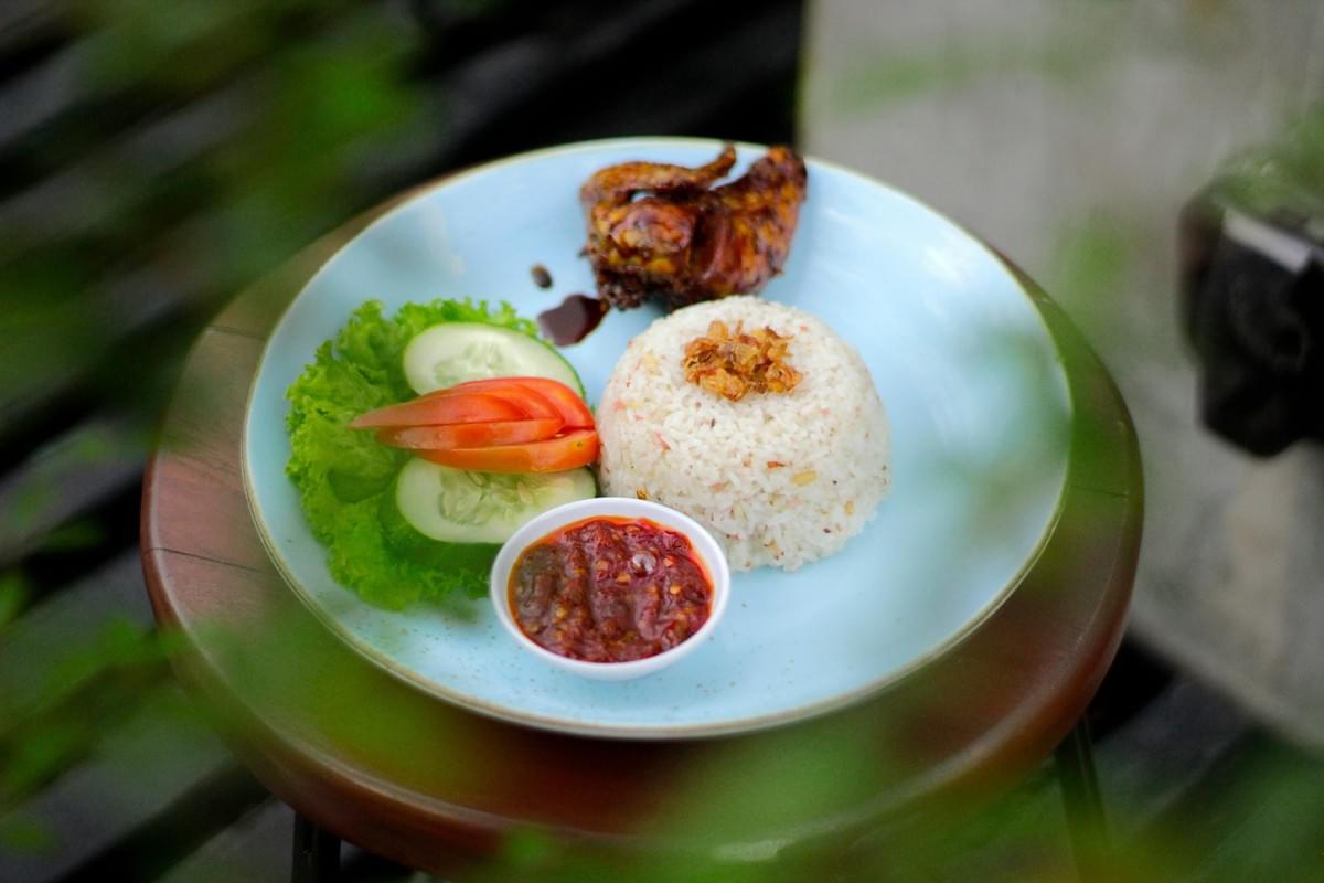 A plate of roasted chicken served with a dome of rice topped with crispy fried shallots is set on a round wooden stool. It includes a side garnish of fresh lettuce, cucumber slices, tomato wedges, and a small dish of red chili paste. A plate of roasted chicken served with a dome of rice topped with crispy fried shallots is set on a round wooden stool. It includes a side garnish of fresh lettuce, cucumber slices, tomato wedges, and a small dish of red chili paste.