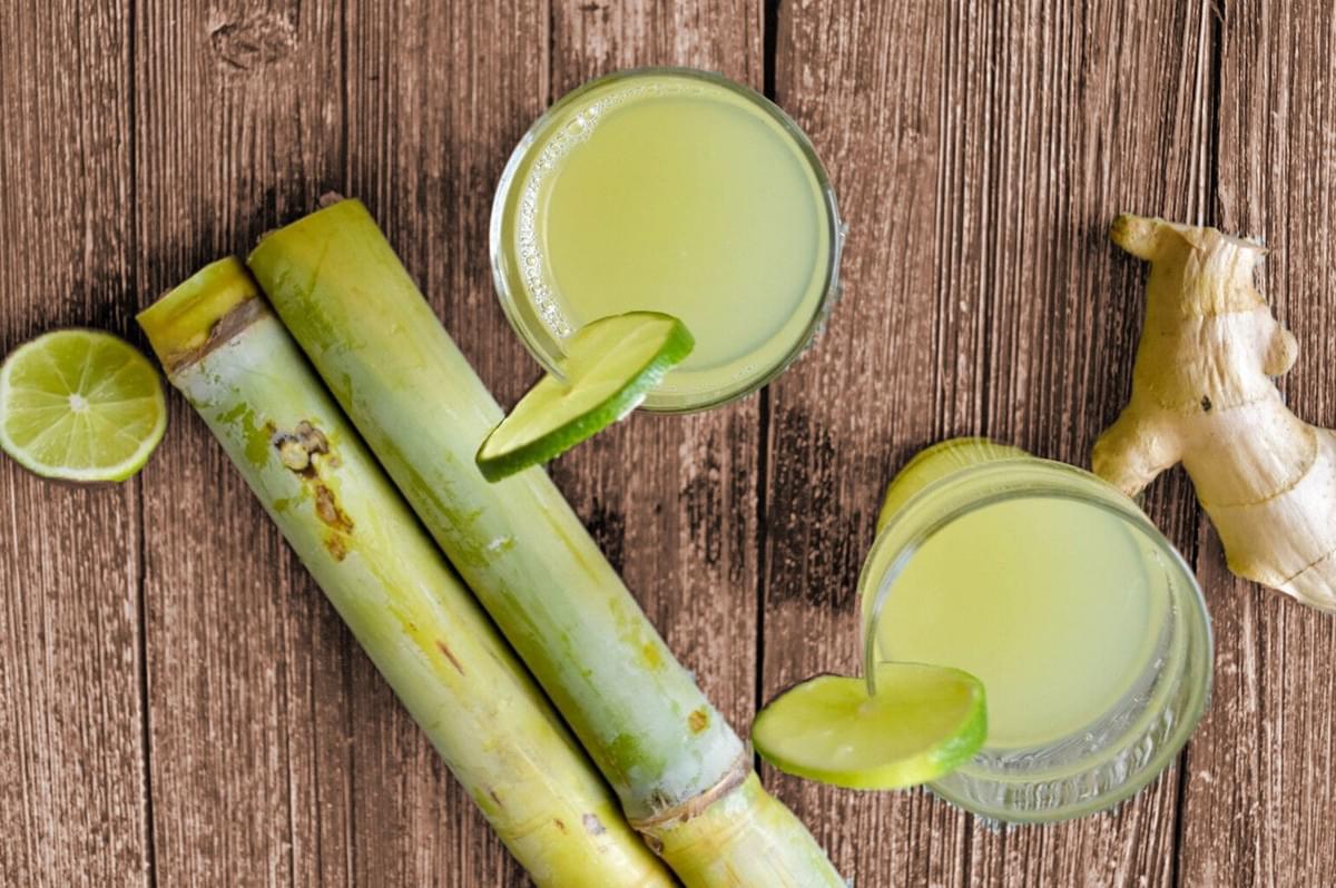 Two glasses of sugarcane juice garnished with lime slices, surrounded by sugarcane stalks, a lime half, and ginger on a rustic wooden table. Refreshing vibe. Two glasses of sugarcane juice garnished with lime slices, surrounded by sugarcane stalks, a lime half, and ginger on a rustic wooden table. Refreshing vibe.