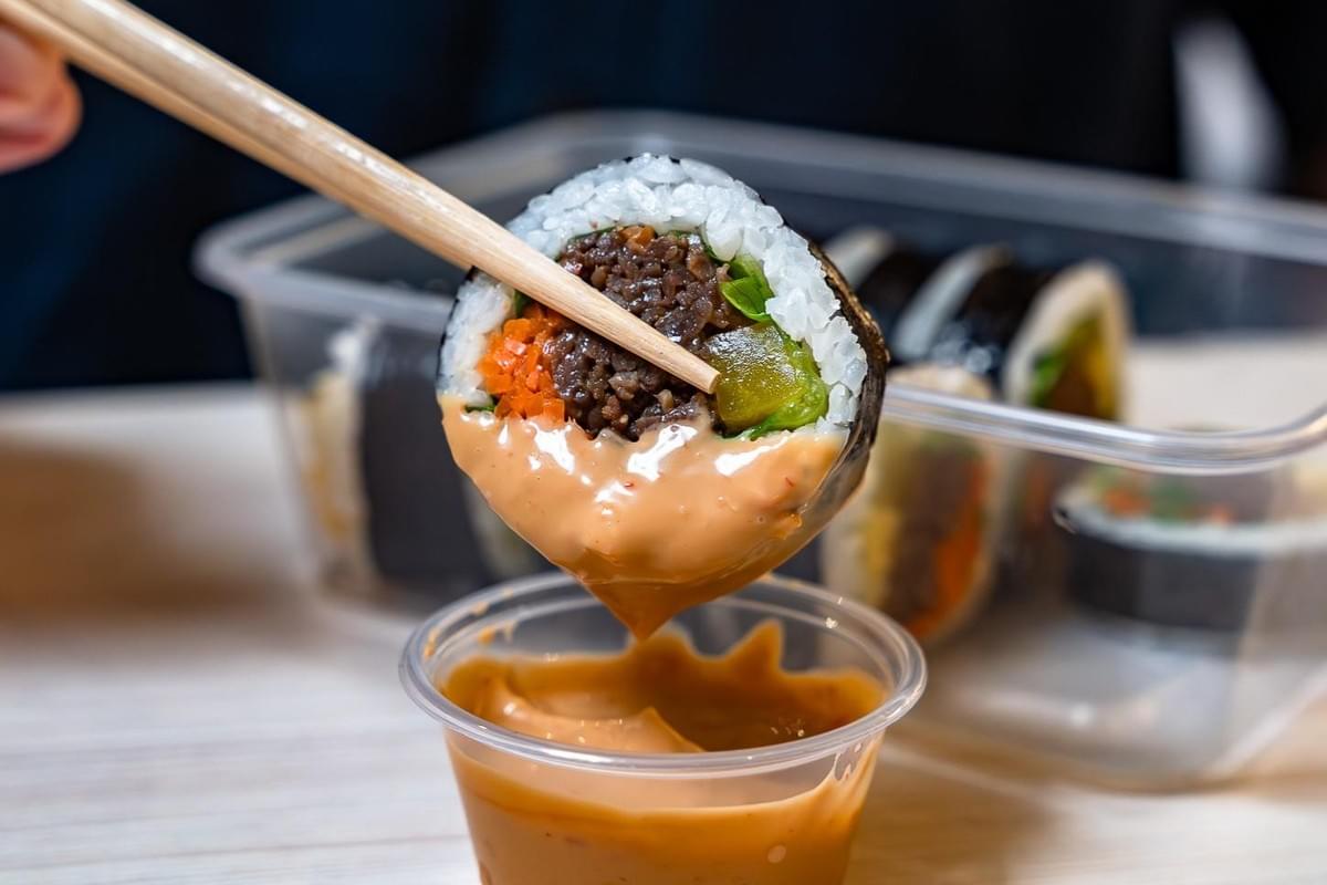 Wooden chopsticks hold a piece of gimbap filled with seasoned meat, rice, and vegetables over a small cup of creamy sauce. The roll is partially coated in the savory dip, with a clear takeout container of remaining gimbap slices sitting in the blurred background. Wooden chopsticks hold a piece of gimbap filled with seasoned meat, rice, and vegetables over a small cup of creamy sauce. The roll is partially coated in the savory dip, with a clear takeout container of remaining gimbap slices sitting in the blurred background.