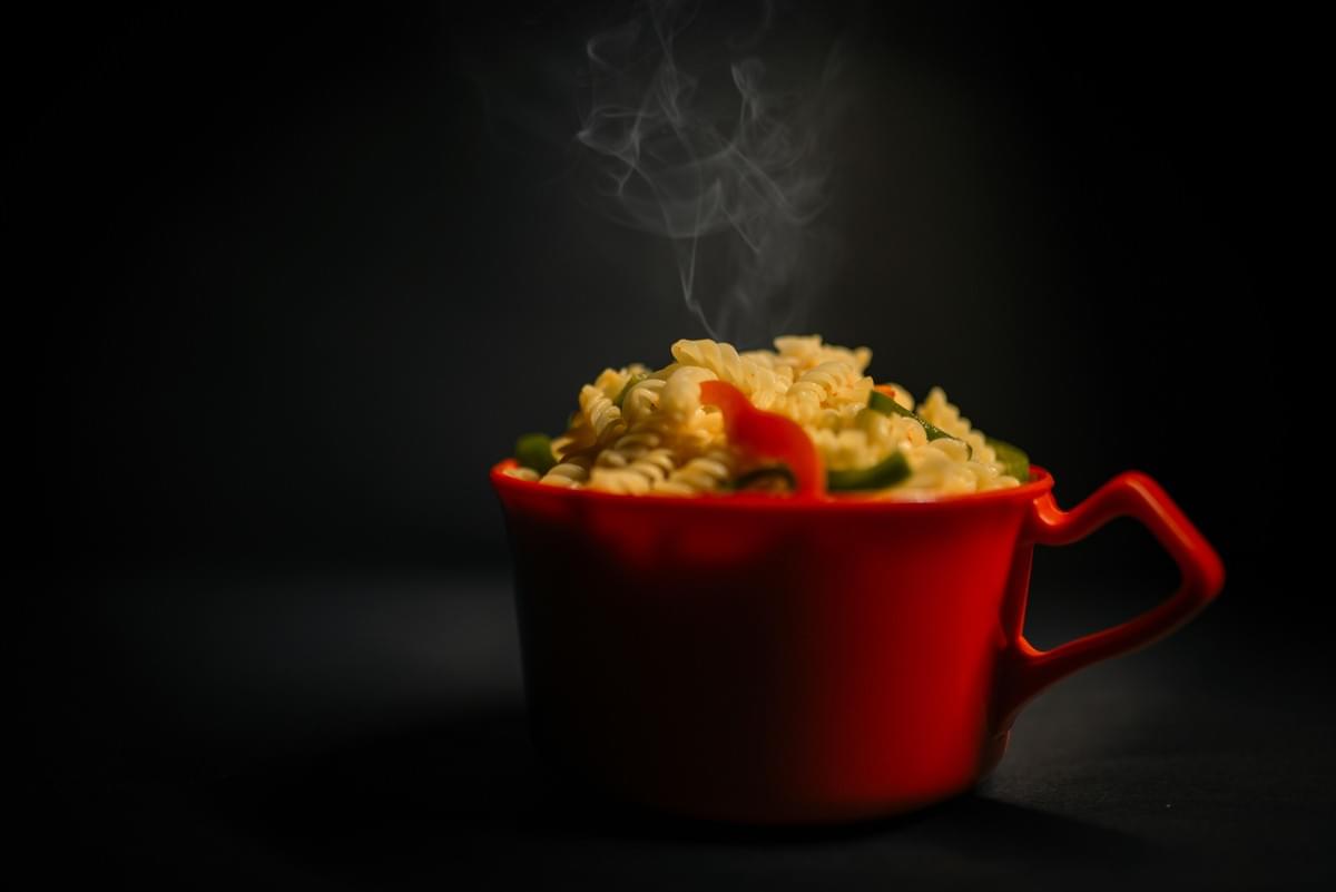 A bright red mug is filled to the brim with hot spiral pasta mixed with sliced red and green peppers. Visible steam swirls upward from the freshly prepared dish against a dark background. A bright red mug is filled to the brim with hot spiral pasta mixed with sliced red and green peppers. Visible steam swirls upward from the freshly prepared dish against a dark background.
