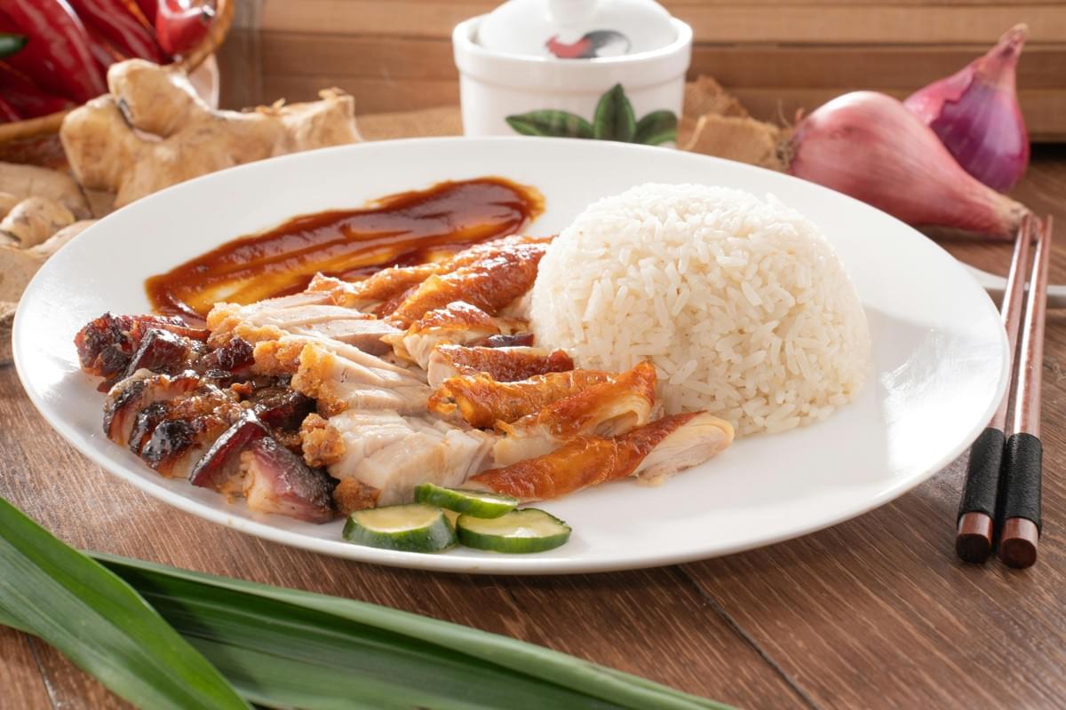 The image shows a white plate of roasted meat rice featuring a dome of white rice, sliced roasted pork belly, char siu, and roasted chicken drizzled with a dark savory sauce. The dish is garnished with sliced cucumbers and set against a rustic wooden background with fresh ingredients like ginger, red onions, and pandan leaves. The image shows a white plate of roasted meat rice featuring a dome of white rice, sliced roasted pork belly, char siu, and roasted chicken drizzled with a dark savory sauce. The dish is garnished with sliced cucumbers and set against a rustic wooden background with fresh ingredients like ginger, red onions, and pandan leaves.