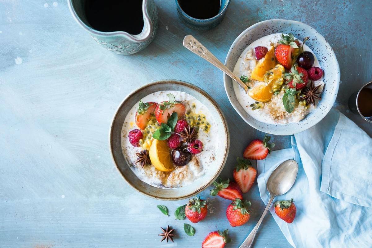 The image shows two bowls of porridge topped with vibrant fresh fruits like strawberries, raspberries, and citrus slices. Set against a textured blue background, the scene also features whole strawberries, star anise, and a pitcher of dark liquid. The image shows two bowls of porridge topped with vibrant fresh fruits like strawberries, raspberries, and citrus slices. Set against a textured blue background, the scene also features whole strawberries, star anise, and a pitcher of dark liquid.