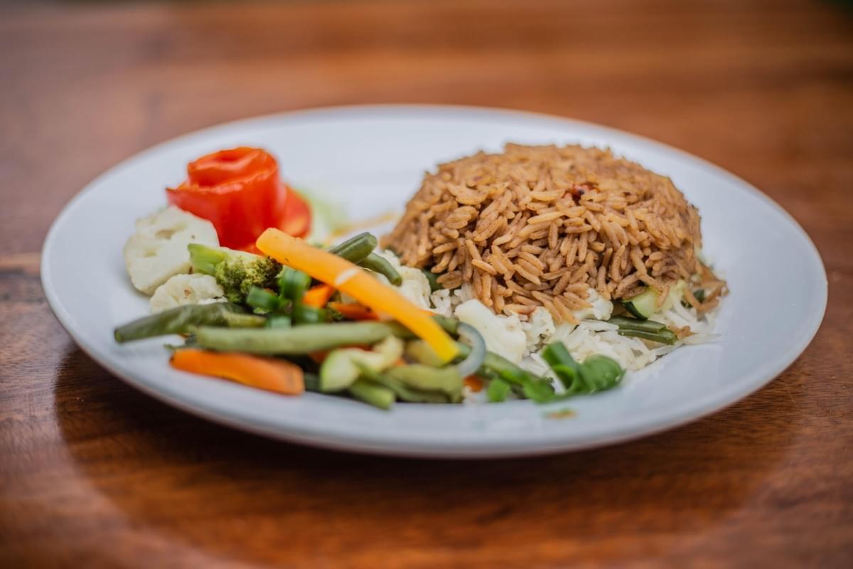 A white plate sits on a wooden surface, featuring a mound of seasoned brown rice served alongside a bed of white rice. The meal is accompanied by an assortment of mixed vegetables, including carrots, green beans, broccoli, cauliflower, and a decorative red tomato slice. A white plate sits on a wooden surface, featuring a mound of seasoned brown rice served alongside a bed of white rice. The meal is accompanied by an assortment of mixed vegetables, including carrots, green beans, broccoli, cauliflower, and a decorative red tomato slice.