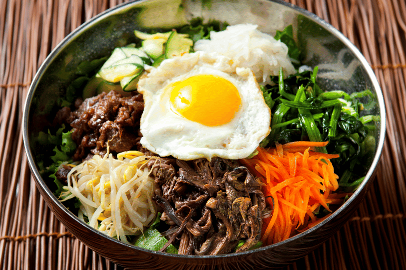 A metal bowl filled with bibimbap featuring distinct sections of spinach, julienned carrots, marinated ferns (gosari), bean sprouts, and beef. A perfectly round sunny-side-up egg sits in the center against a woven bamboo mat background. A metal bowl filled with bibimbap featuring distinct sections of spinach, julienned carrots, marinated ferns (gosari), bean sprouts, and beef. A perfectly round sunny-side-up egg sits in the center against a woven bamboo mat background.