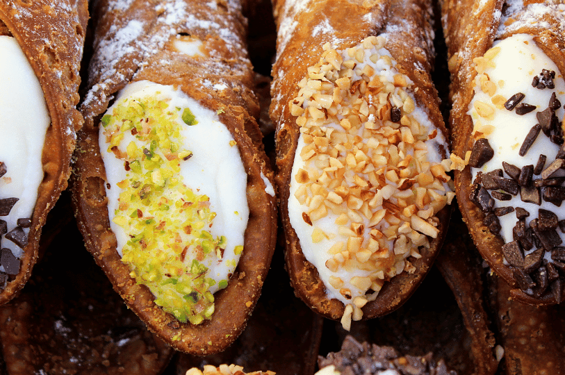 An extreme close-up of three cannoli ends showcasing different toppings: vibrant green crushed pistachios, chopped toasted almonds, and dark chocolate chips embedded in the ricotta filling. An extreme close-up of three cannoli ends showcasing different toppings: vibrant green crushed pistachios, chopped toasted almonds, and dark chocolate chips embedded in the ricotta filling.