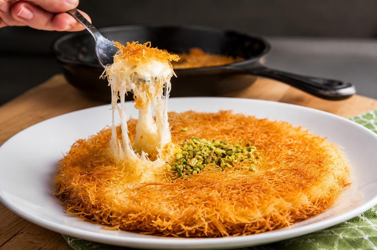 A fork lifting a bite of crispy, golden kunafa dessert to create a long, stretchy cheese pull from a round pastry topped with chopped pistachios on a white plate. A fork lifting a bite of crispy, golden kunafa dessert to create a long, stretchy cheese pull from a round pastry topped with chopped pistachios on a white plate.