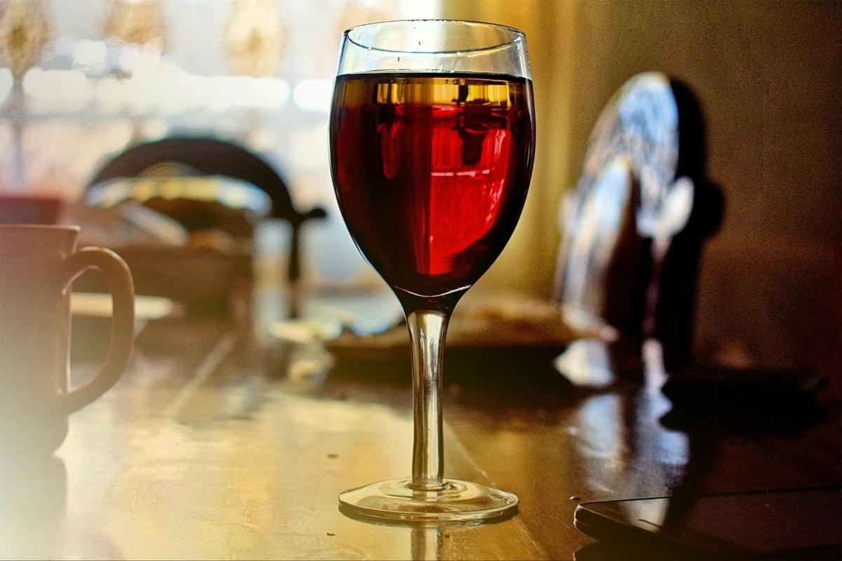 In the image, a wine glass filled with a deep red liquid stands prominently on a polished wooden table. The warm, dimly lit background features blurred shapes of chairs and a mug, creating a cozy and intimate atmosphere. In the image, a wine glass filled with a deep red liquid stands prominently on a polished wooden table. The warm, dimly lit background features blurred shapes of chairs and a mug, creating a cozy and intimate atmosphere.