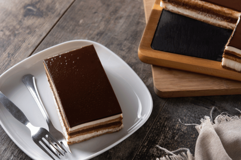 An overhead view of a pristine slice of opera cake with a glossy dark chocolate top on a white plate, set on a rustic wooden table alongside two silver forks and a serving board with more cake slices. An overhead view of a pristine slice of opera cake with a glossy dark chocolate top on a white plate, set on a rustic wooden table alongside two silver forks and a serving board with more cake slices.