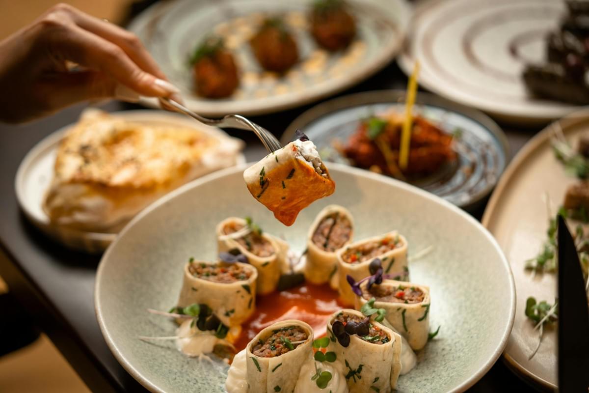 A hand uses a fork to lift a piece of savory rolled wrap from a plate filled with similar sliced pinwheels arranged over a red sauce and white cream. In the background, other gourmet dishes are subtly blurred on a dark restaurant table. A hand uses a fork to lift a piece of savory rolled wrap from a plate filled with similar sliced pinwheels arranged over a red sauce and white cream. In the background, other gourmet dishes are subtly blurred on a dark restaurant table.