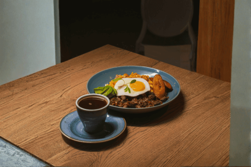 An artistic, high-angle shot of a Bandeja Paisa served in a blue ceramic bowl next to a cup of Colombian black coffee, emphasizing the egg, plantain, and bean base. An artistic, high-angle shot of a Bandeja Paisa served in a blue ceramic bowl next to a cup of Colombian black coffee, emphasizing the egg, plantain, and bean base.