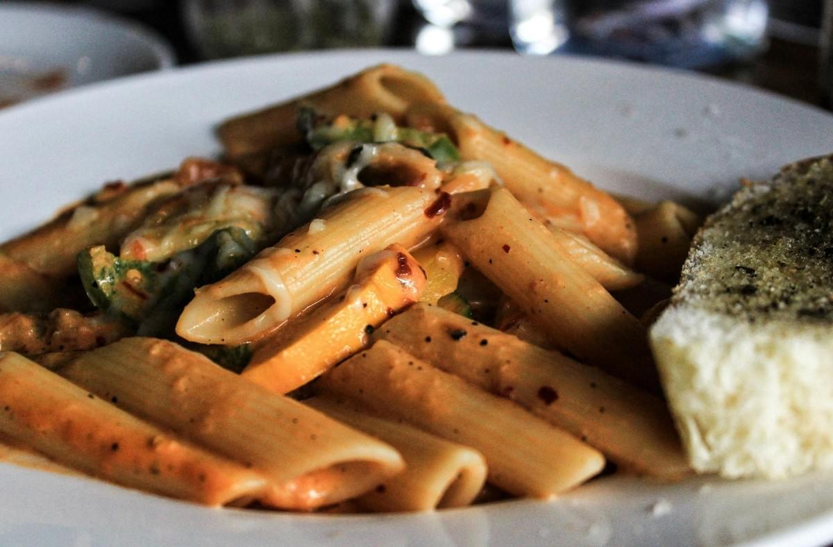 A plate of penne pasta is coated in a creamy tomato sauce and mixed with vegetables and melted cheese. A slice of seasoned garlic bread rests on the side of the white dish. A plate of penne pasta is coated in a creamy tomato sauce and mixed with vegetables and melted cheese. A slice of seasoned garlic bread rests on the side of the white dish.