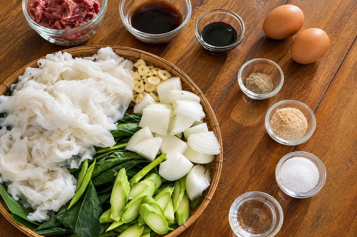 Overhead shot of cooking ingredients including wide rice noodles, chopped onions, greens, raw beef, eggs, and seasonings arrayed on a wooden surface. Overhead shot of cooking ingredients including wide rice noodles, chopped onions, greens, raw beef, eggs, and seasonings arrayed on a wooden surface.