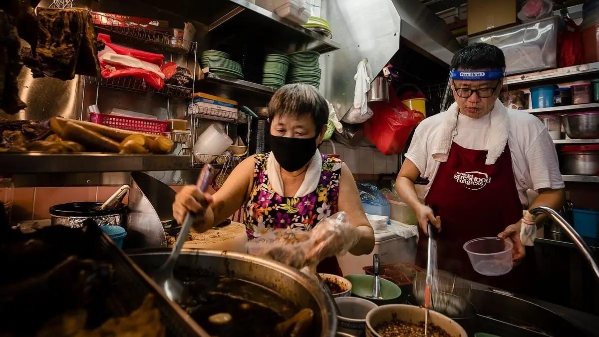 Inside a bustling kitchen, two vendors wearing protective masks and face shields work diligently to prepare food from large steaming pots and organized shelves of ingredients. Inside a bustling kitchen, two vendors wearing protective masks and face shields work diligently to prepare food from large steaming pots and organized shelves of ingredients.