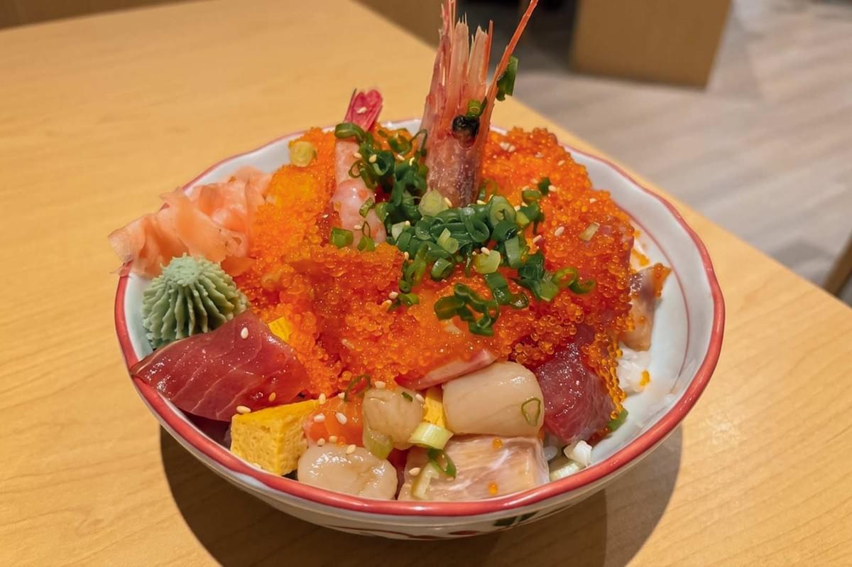 This close-up shot highlights a generously loaded chirashi don bowl served on a light wooden table, overflowing with a colorful variety of diced fresh sashimi, including tuna, scallops, and tamago cubes. The dish is covered with a thick mound of bright orange tobiko fish roe, topped with a whole sweet prawn featuring its head standing upright, sprinkled with chopped scallions, and garnished on the side with pickled ginger and a piped mound of wasabi. This close-up shot highlights a generously loaded chirashi don bowl served on a light wooden table, overflowing with a colorful variety of diced fresh sashimi, including tuna, scallops, and tamago cubes. The dish is covered with a thick mound of bright orange tobiko fish roe, topped with a whole sweet prawn featuring its head standing upright, sprinkled with chopped scallions, and garnished on the side with pickled ginger and a piped mound of wasabi.