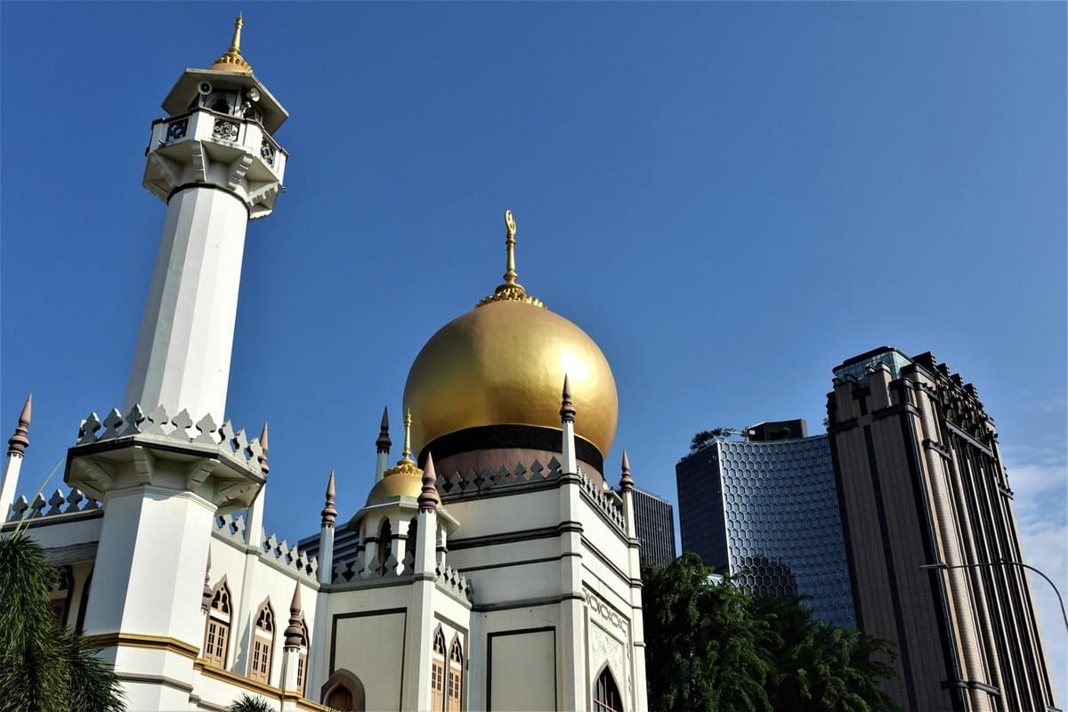 A low-angle view highlights a white mosque featuring a prominent golden dome and a tall minaret set against a clear blue sky. Modern skyscrapers rise in the background behind the traditional architectural structure. A low-angle view highlights a white mosque featuring a prominent golden dome and a tall minaret set against a clear blue sky. Modern skyscrapers rise in the background behind the traditional architectural structure.