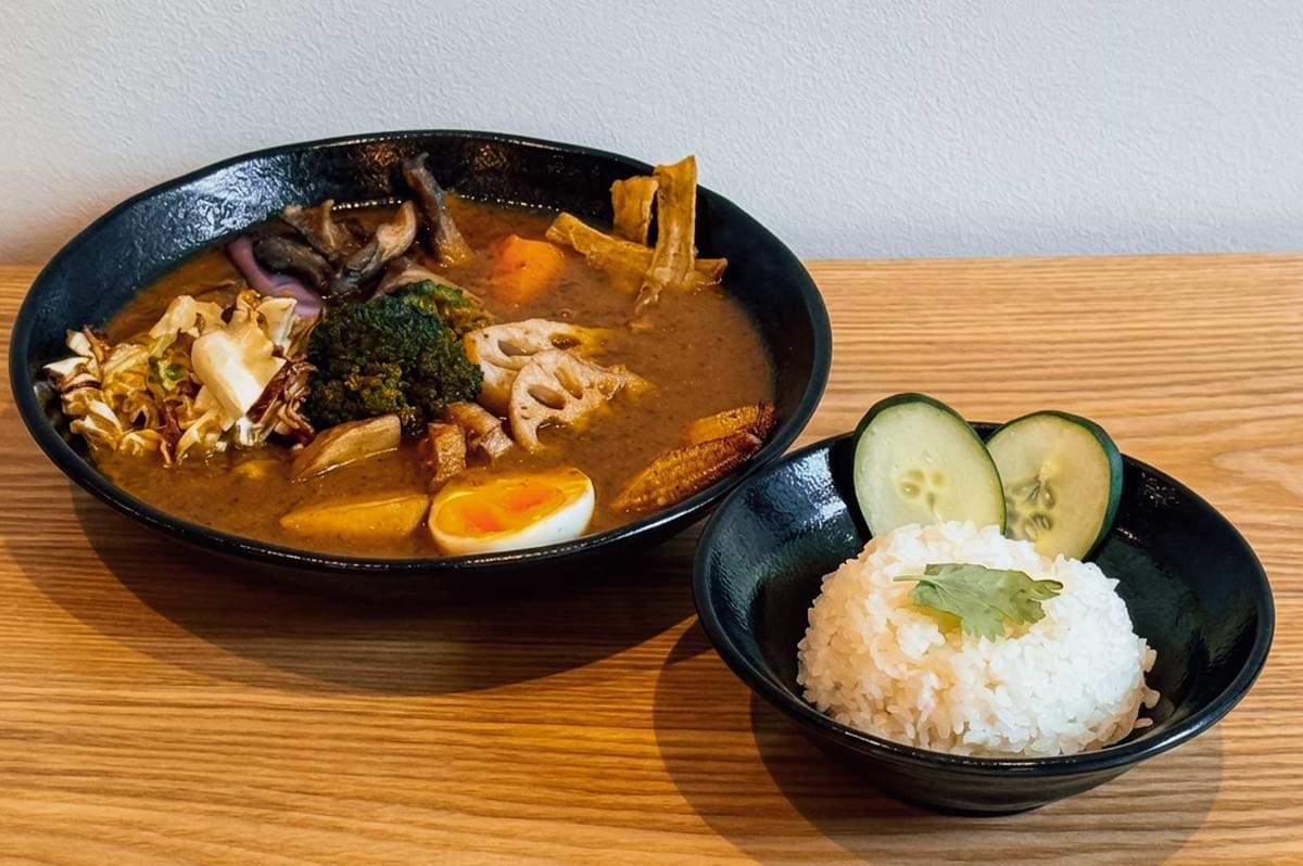 A serving of Japanese soup curry from "Soup Curry By Ki-setsu" in Singapore, presented on a natural wood surface alongside a small black bowl of white rice garnished with fresh cilantro and cucumber slices, featuring a fragrant curry broth packed with a soft-boiled egg half, lotus root, broccoli, sauteed mushrooms, shredded cabbage, baby corn, and fried burdock root chips. A serving of Japanese soup curry from "Soup Curry By Ki-setsu" in Singapore, presented on a natural wood surface alongside a small black bowl of white rice garnished with fresh cilantro and cucumber slices, featuring a fragrant curry broth packed with a soft-boiled egg half, lotus root, broccoli, sauteed mushrooms, shredded cabbage, baby corn, and fried burdock root chips.