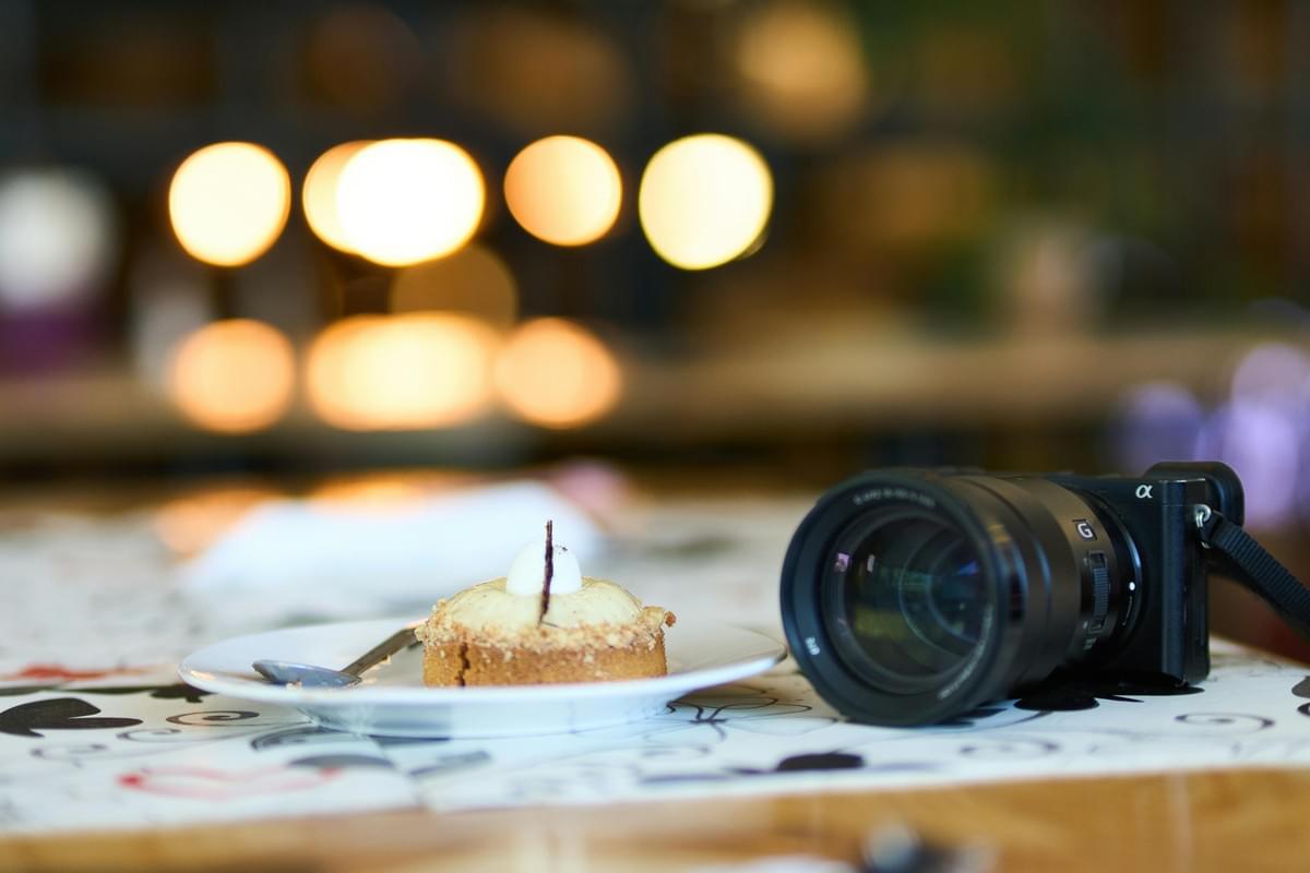 A small, elegant pastry sits on a white plate alongside a spoon on a patterned tablecloth. To its right, a black mirrorless camera with a large lens is positioned on the table against a warm, blurred background of bokeh lights. A small, elegant pastry sits on a white plate alongside a spoon on a patterned tablecloth. To its right, a black mirrorless camera with a large lens is positioned on the table against a warm, blurred background of bokeh lights.