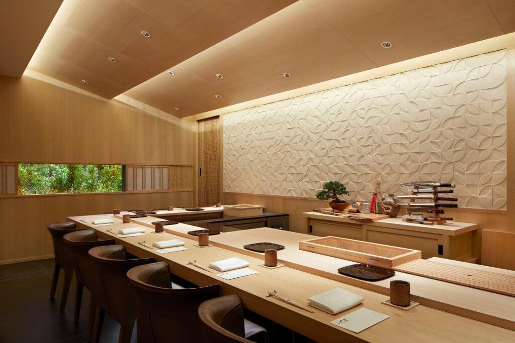 An elegant, modern Japanese dining room featuring a U-shaped light wood counter lined with dark leather chairs and set for a meal. Warm ambient lighting accents a textured pattern wall, wood paneling, and a chef's station displayed with traditional knives and a bonsai tree. An elegant, modern Japanese dining room featuring a U-shaped light wood counter lined with dark leather chairs and set for a meal. Warm ambient lighting accents a textured pattern wall, wood paneling, and a chef's station displayed with traditional knives and a bonsai tree.