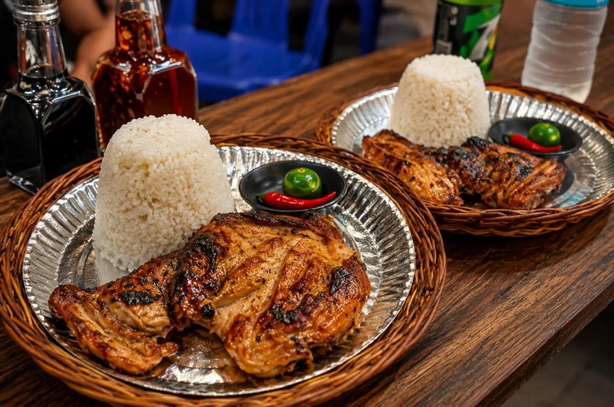 "Two woven tray meals of grilled chicken, molded rice, calamansi, and chili served on a wooden table.” "Two woven tray meals of grilled chicken, molded rice, calamansi, and chili served on a wooden table.”