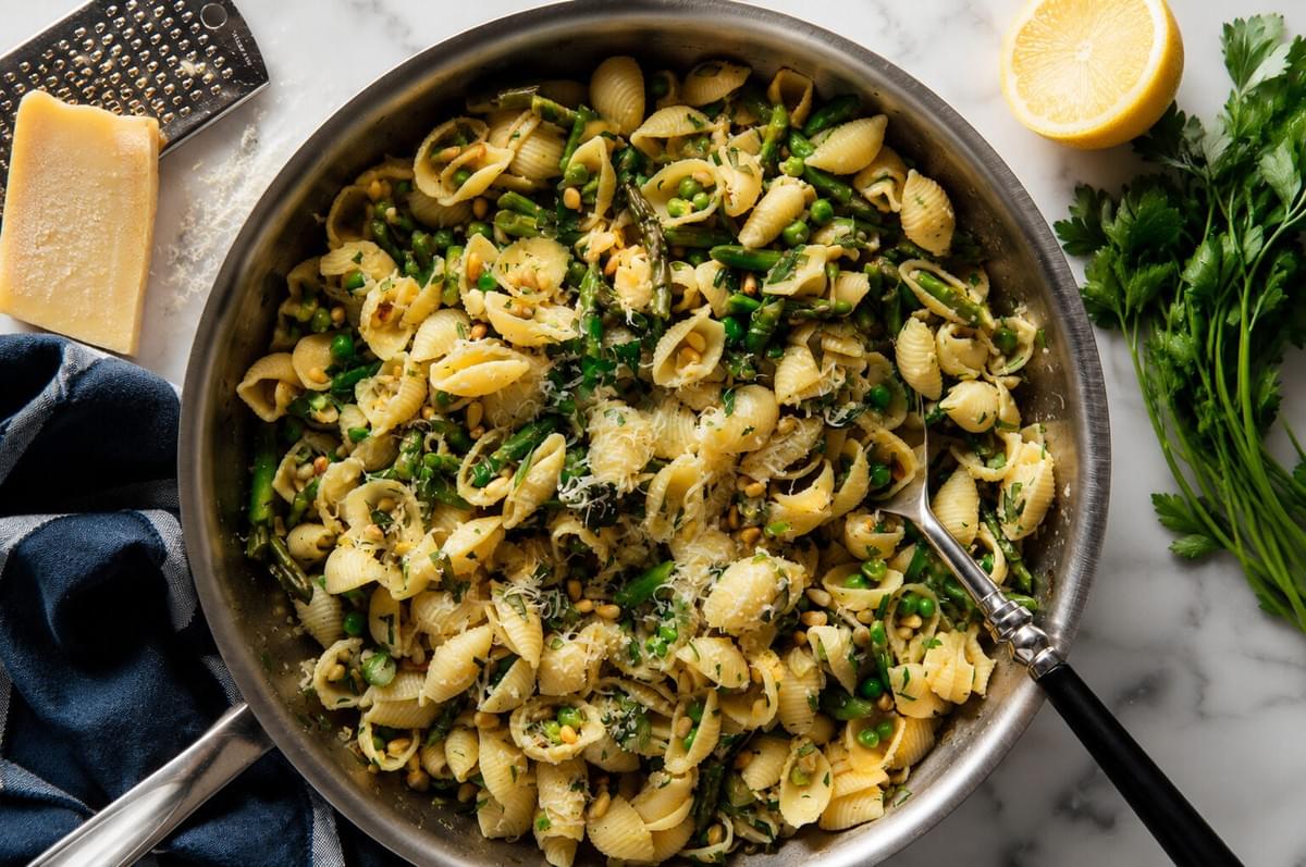 Overhead view of shell pasta with peas, asparagus, and herbs in a large skillet alongside parmesan cheese and a lemon half. Overhead view of shell pasta with peas, asparagus, and herbs in a large skillet alongside parmesan cheese and a lemon half.