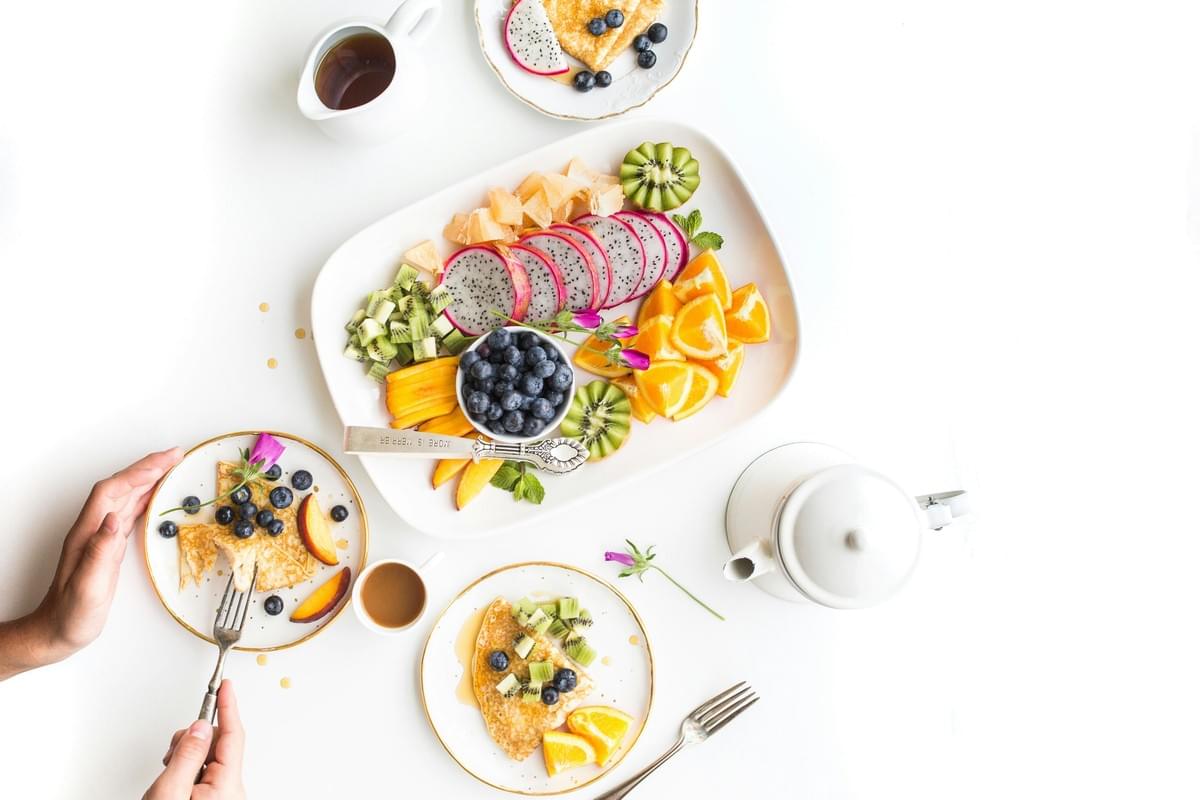The image displays a vibrant breakfast spread featuring a large platter of sliced tropical fruits, including dragon fruit, kiwi, and oranges. Small plates with crepes topped with fresh berries and kiwi accompany the fruit platter on a clean white surface. The image displays a vibrant breakfast spread featuring a large platter of sliced tropical fruits, including dragon fruit, kiwi, and oranges. Small plates with crepes topped with fresh berries and kiwi accompany the fruit platter on a clean white surface.
