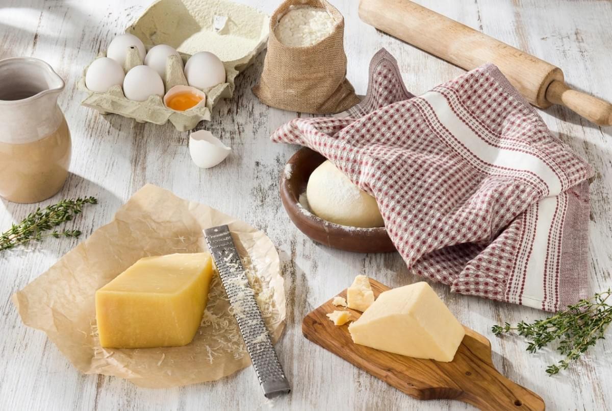 Baking ingredients layout featuring blocks of cheese, a Microplane grater, eggs, a sack of flour, dough in a bowl, and a rolling pin. Baking ingredients layout featuring blocks of cheese, a Microplane grater, eggs, a sack of flour, dough in a bowl, and a rolling pin.