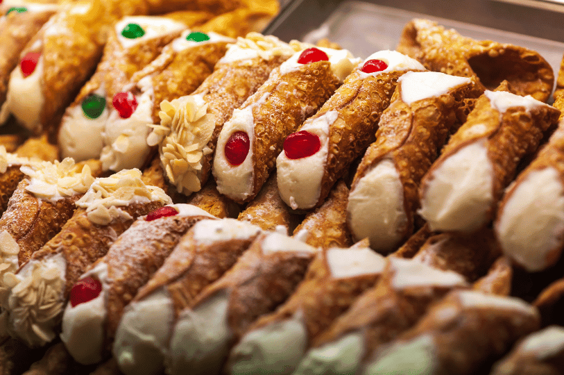 A bakery display case featuring rows of cannoli with diverse toppings, including candied red and green cherries, sliced almonds, and plain ricotta cream. A bakery display case featuring rows of cannoli with diverse toppings, including candied red and green cherries, sliced almonds, and plain ricotta cream.