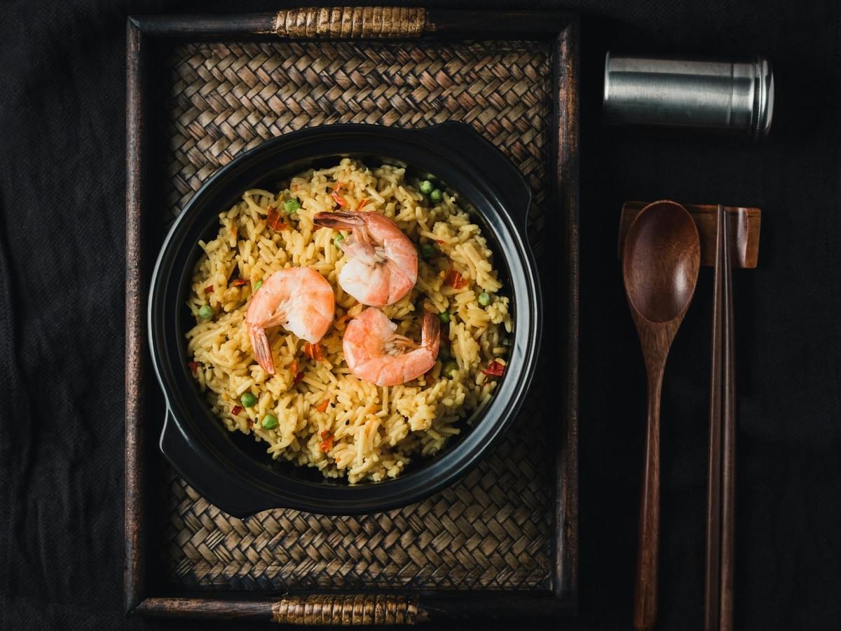 A top-down view shows a black ceramic bowl filled with seasoned rice and topped with three cooked shrimp, set inside a woven wooden tray. Beside the bowl, a wooden spoon, chopsticks, and a small metal shaker rest on a dark backdrop. A top-down view shows a black ceramic bowl filled with seasoned rice and topped with three cooked shrimp, set inside a woven wooden tray. Beside the bowl, a wooden spoon, chopsticks, and a small metal shaker rest on a dark backdrop.