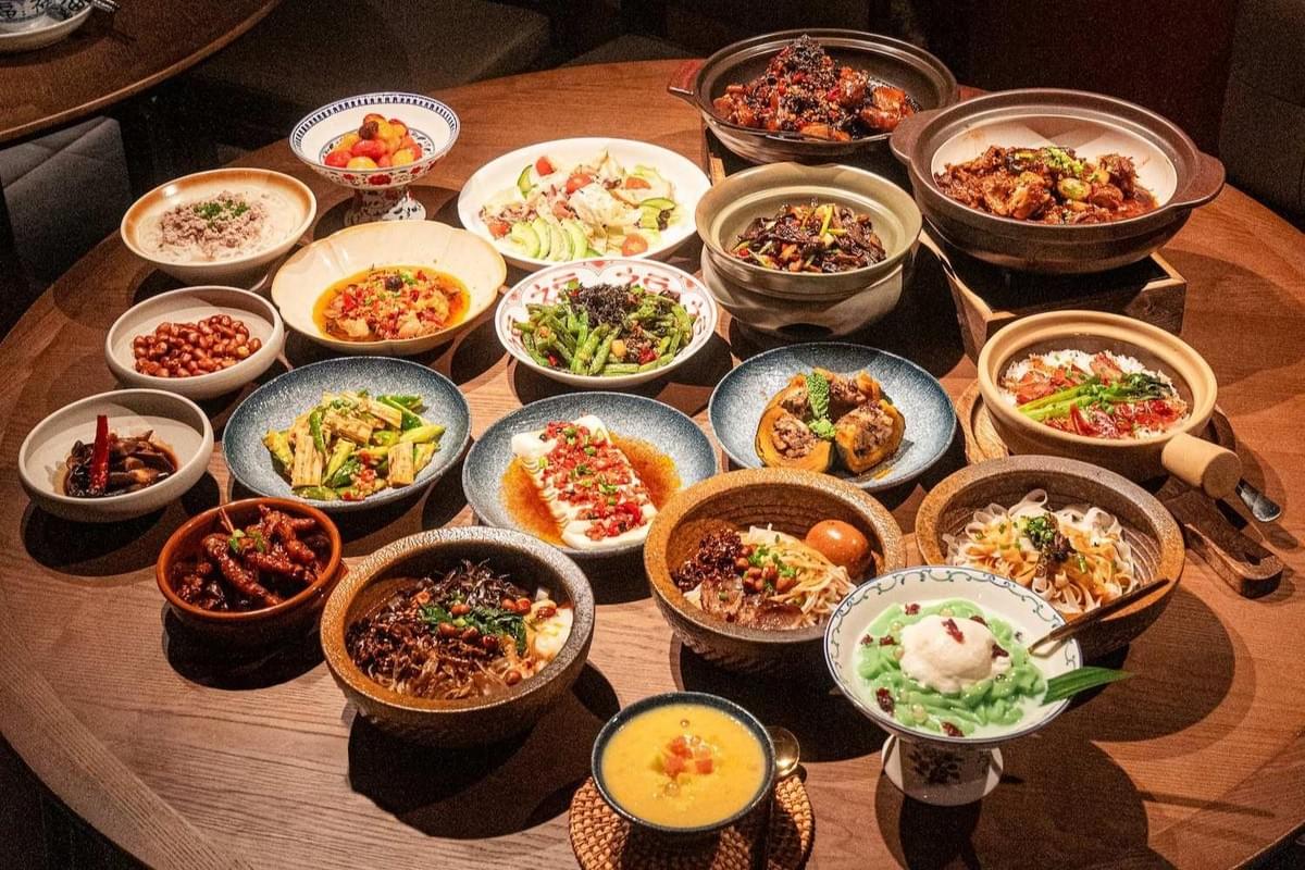 A large round wooden table is filled with an elaborate spread of various Chinese dishes served in clay pots, ceramic bowls, and colorful plates. The colorful family-style meal features a wide variety of stir-fries, noodle dishes, steamed tofu with chili sauce, vegetables, soup, and iced desserts. A large round wooden table is filled with an elaborate spread of various Chinese dishes served in clay pots, ceramic bowls, and colorful plates. The colorful family-style meal features a wide variety of stir-fries, noodle dishes, steamed tofu with chili sauce, vegetables, soup, and iced desserts.