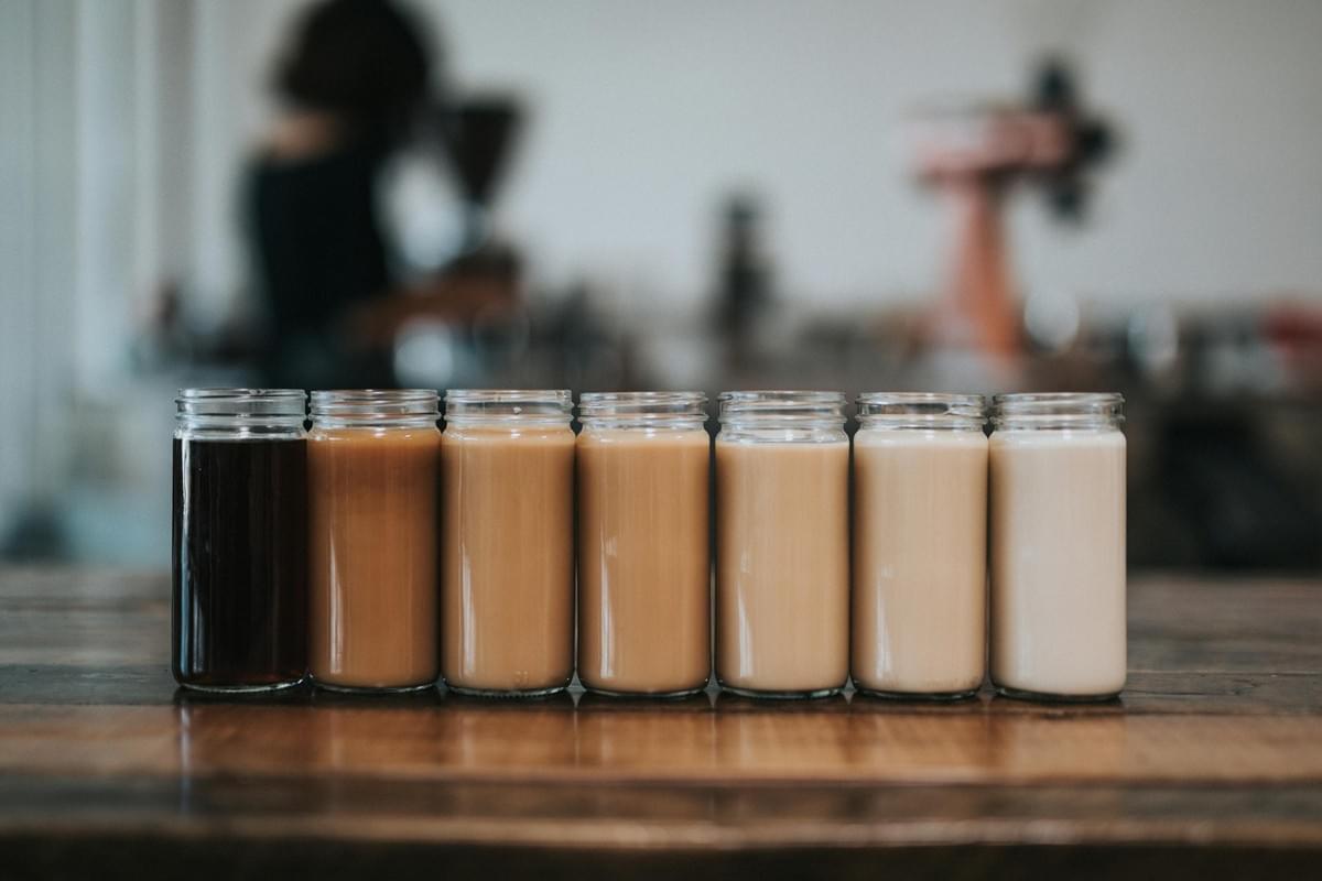 Seven glass jars filled with coffee beverages sit in a neat row on a wooden table, ranging in shade from deep black on the left to pale cream on the right. In the softly blurred background, a barista works inside a brightly lit coffee shop setting. Seven glass jars filled with coffee beverages sit in a neat row on a wooden table, ranging in shade from deep black on the left to pale cream on the right. In the softly blurred background, a barista works inside a brightly lit coffee shop setting.