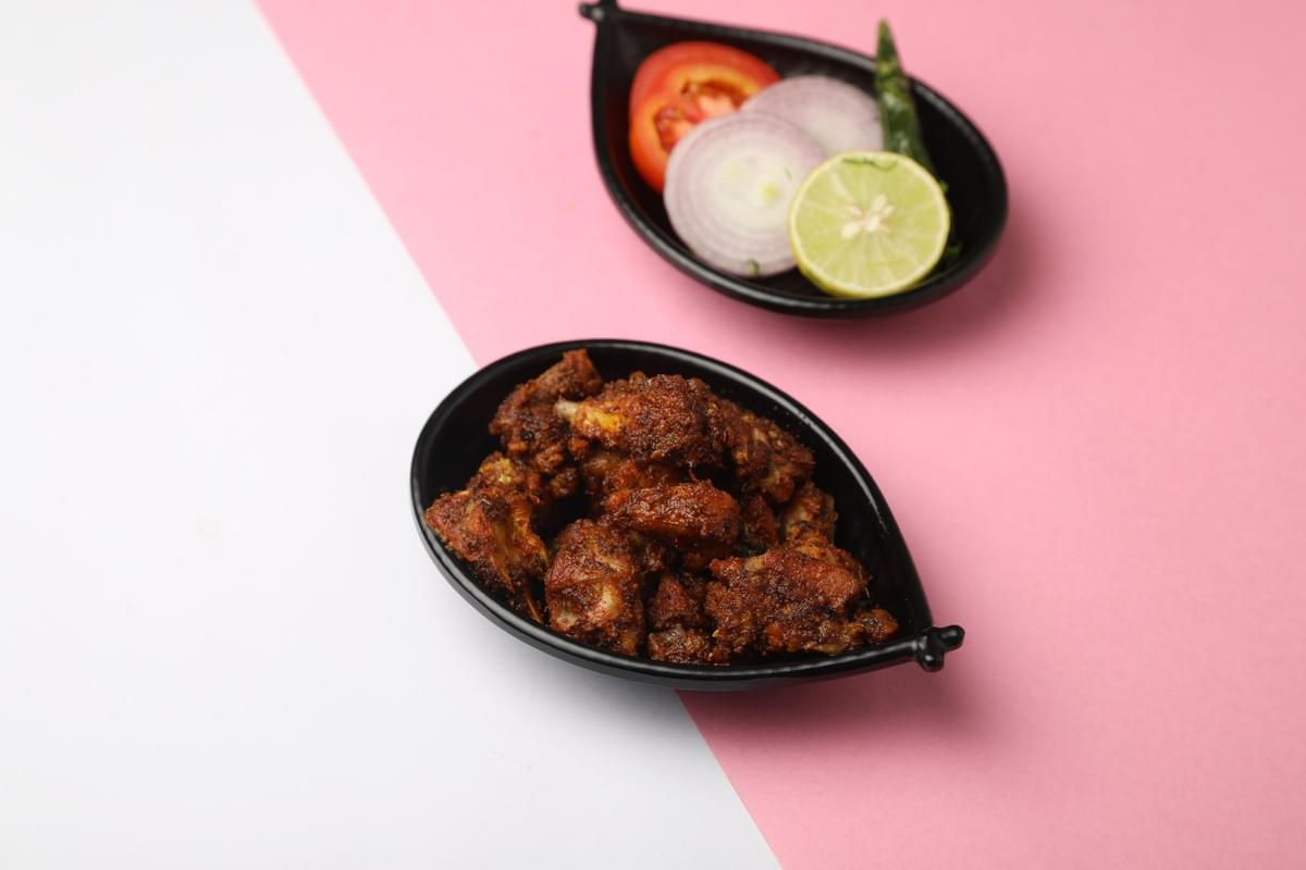 The image features a black leaf-shaped bowl filled with roasted or fried spiced chicken pieces, placed on a diagonally split white and pink background. In the upper right background, a matching black dish contains a side garnish consisting of sliced red onions, a tomato slice, a lime wedge, and a green chili pepper. The image features a black leaf-shaped bowl filled with roasted or fried spiced chicken pieces, placed on a diagonally split white and pink background. In the upper right background, a matching black dish contains a side garnish consisting of sliced red onions, a tomato slice, a lime wedge, and a green chili pepper.