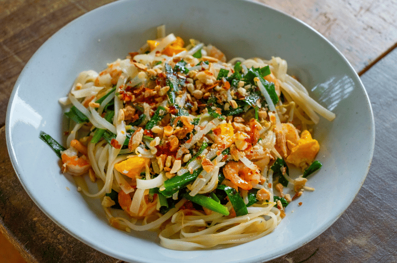 A close-up bowl of Pad Thai featuring flat rice noodles, fresh green chives, bean sprouts, succulent shrimp, and a generous topping of crushed peanuts and chili flakes. A close-up bowl of Pad Thai featuring flat rice noodles, fresh green chives, bean sprouts, succulent shrimp, and a generous topping of crushed peanuts and chili flakes.