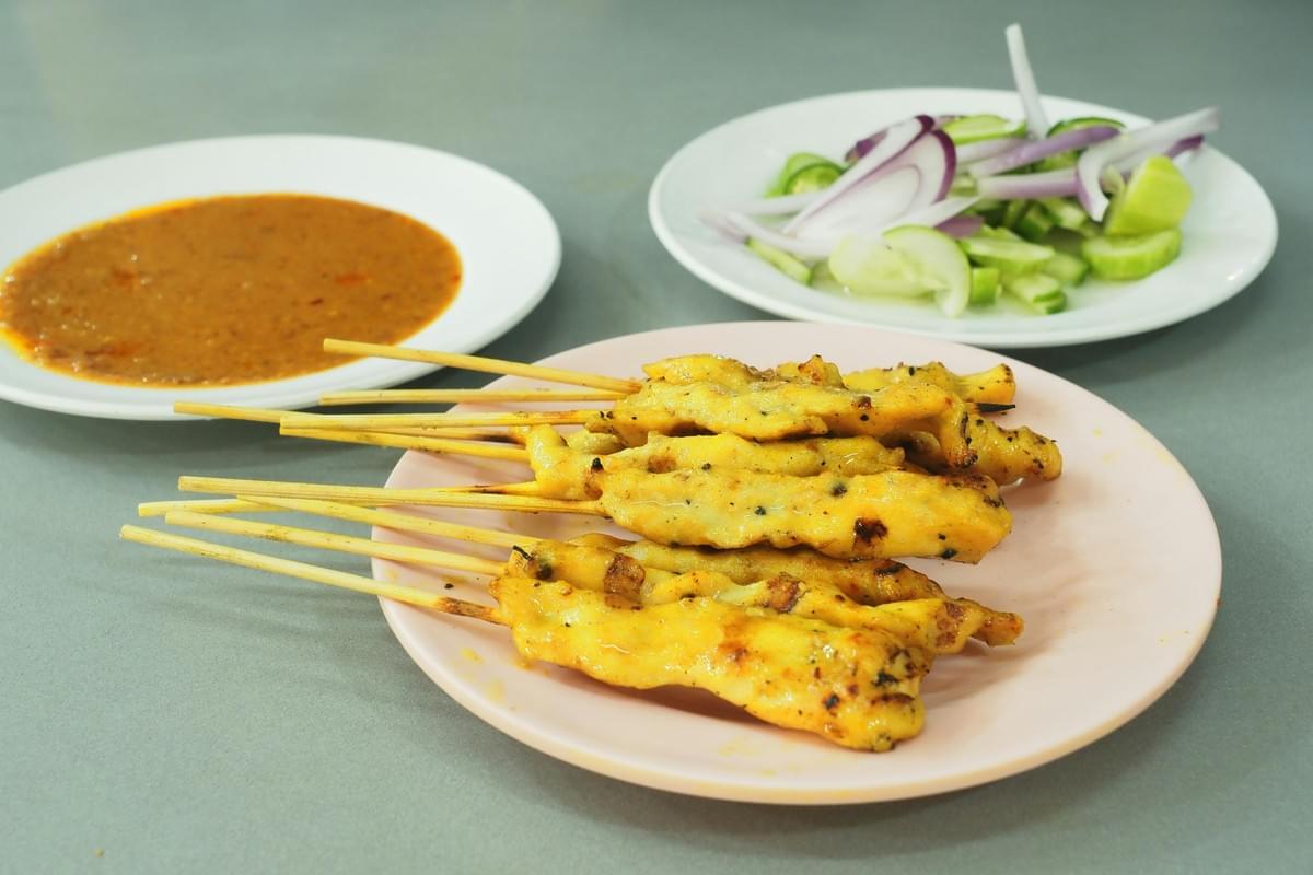 A light pink plate holds several grilled chicken satay skewers stacked together in the foreground. Beside the main dish are two white plates containing a thick peanut dipping sauce and a fresh side of sliced cucumbers and red onions. A light pink plate holds several grilled chicken satay skewers stacked together in the foreground. Beside the main dish are two white plates containing a thick peanut dipping sauce and a fresh side of sliced cucumbers and red onions.