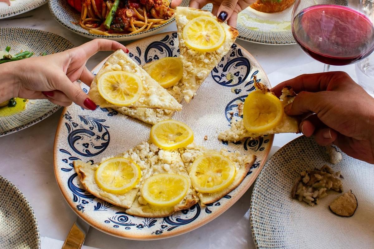 Multiple diners reach simultaneously for slices of a flatbread topped with lemon wheels and crumbled cheese served on a blue-patterned plate. The table is crowded with other dishes, including pasta and risotto, along with a glass of red wine, suggesting a lively shared meal. Multiple diners reach simultaneously for slices of a flatbread topped with lemon wheels and crumbled cheese served on a blue-patterned plate. The table is crowded with other dishes, including pasta and risotto, along with a glass of red wine, suggesting a lively shared meal.