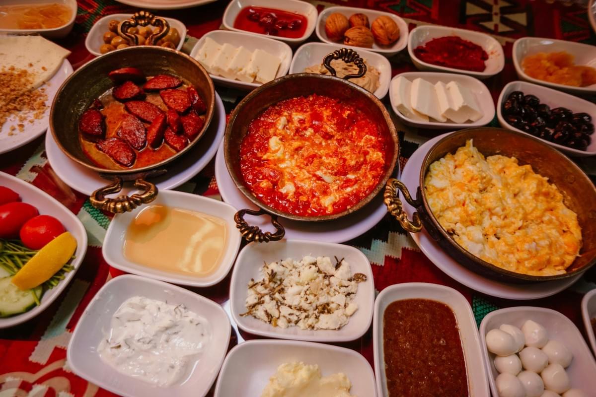 This image captures a lavish traditional Turkish breakfast spread arranged on a patterned red textile, featuring copper pans filled with hot dishes like menemen, scrambled eggs, and spicy sucuk sausage. Surrounding the hot pans is a vast assortment of small white plates holding various cheeses, olives, jams, dips, and fresh vegetables, creating a vibrant and bountiful feast. This image captures a lavish traditional Turkish breakfast spread arranged on a patterned red textile, featuring copper pans filled with hot dishes like menemen, scrambled eggs, and spicy sucuk sausage. Surrounding the hot pans is a vast assortment of small white plates holding various cheeses, olives, jams, dips, and fresh vegetables, creating a vibrant and bountiful feast.