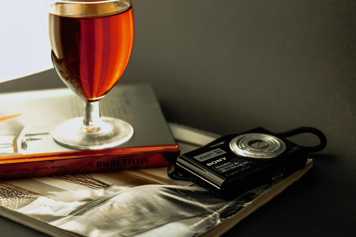 In the image, a stemmed glass filled with an amber-colored liquid rests on a book titled "Small Homes." Beside it, a black Sony Cyber-shot compact digital camera sits on an open magazine under warm, dramatic lighting. In the image, a stemmed glass filled with an amber-colored liquid rests on a book titled "Small Homes." Beside it, a black Sony Cyber-shot compact digital camera sits on an open magazine under warm, dramatic lighting.