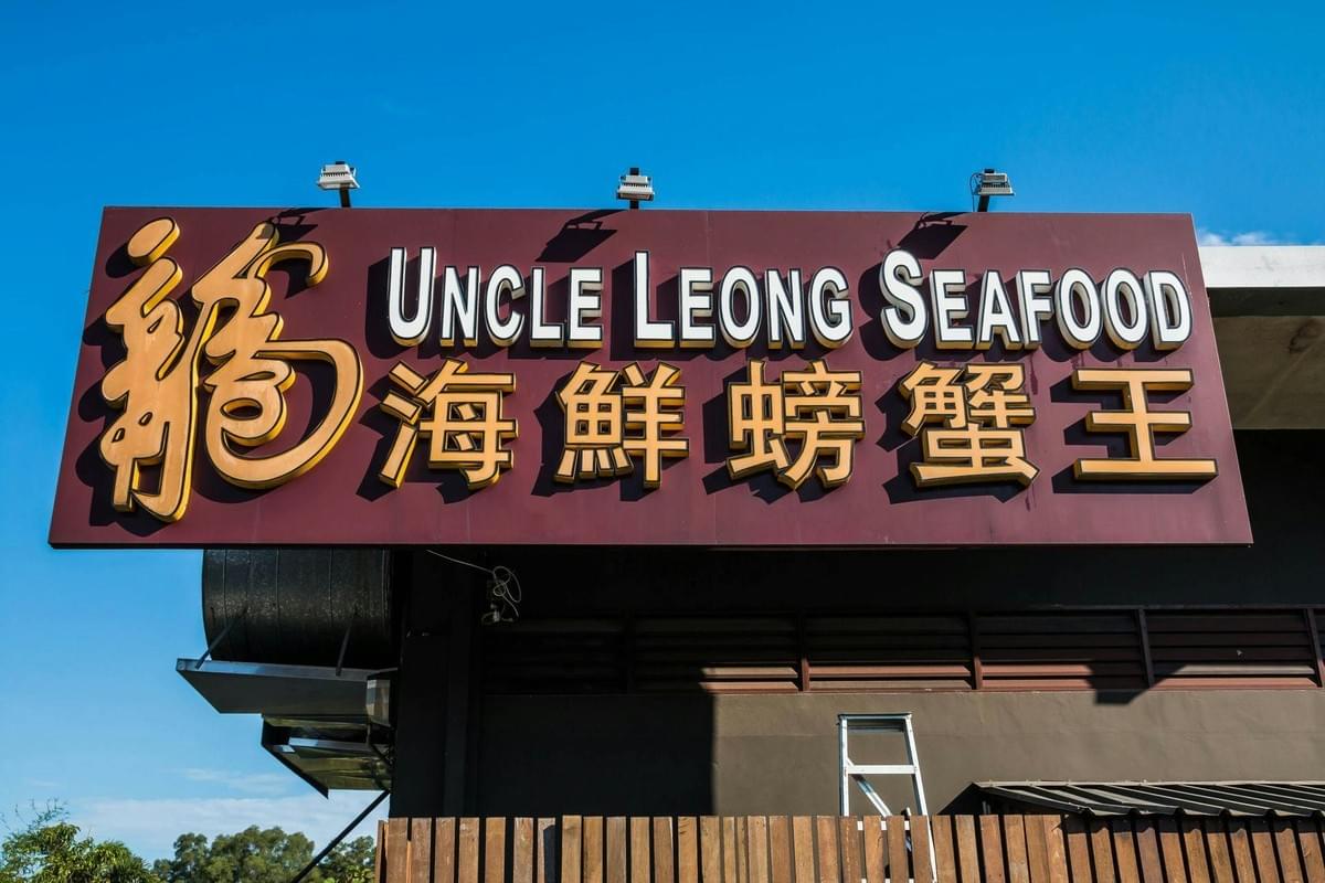 A large brown sign displays the English text "UNCLE LEONG SEAFOOD" alongside prominent gold Chinese characters. The sign is mounted on a dark exterior wall under a clear blue sky. A large brown sign displays the English text "UNCLE LEONG SEAFOOD" alongside prominent gold Chinese characters. The sign is mounted on a dark exterior wall under a clear blue sky.
