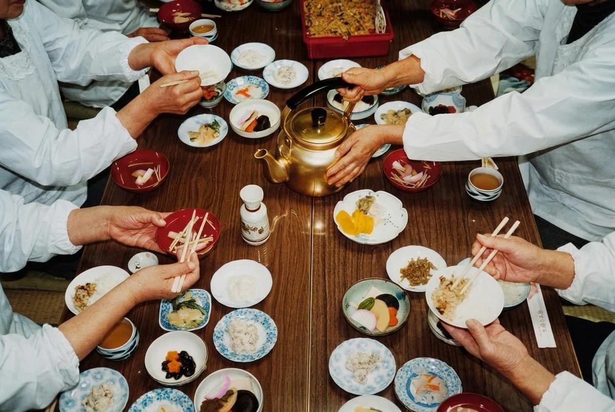 Top-down view of people sitting around a wooden table sharing small bowls of Japanese home-cooked food and tea. Top-down view of people sitting around a wooden table sharing small bowls of Japanese home-cooked food and tea.