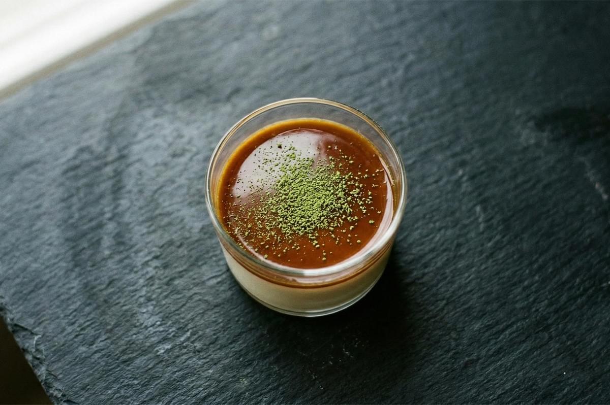 A single glass of creamy white custard topped with a layer of dark caramel and a fine dusting of green matcha powder on a dark stone surface. A single glass of creamy white custard topped with a layer of dark caramel and a fine dusting of green matcha powder on a dark stone surface.