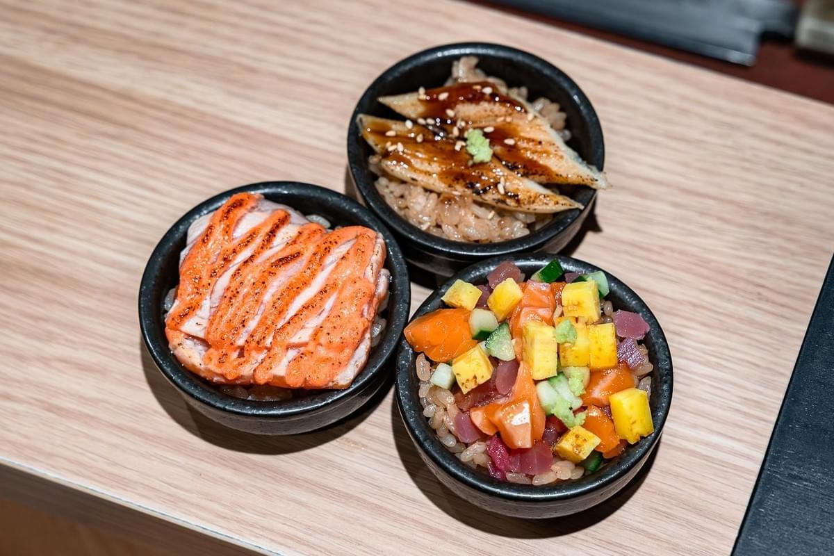 Three mini rice bowls are arranged on a wooden surface, each featuring a distinct Japanese topping over seasoned sushi rice. The toppings include seared salmon drizzled with sauce, glazed eel with sesame seeds, and a colorful mixture of diced sashimi, egg, and cucumber. Three mini rice bowls are arranged on a wooden surface, each featuring a distinct Japanese topping over seasoned sushi rice. The toppings include seared salmon drizzled with sauce, glazed eel with sesame seeds, and a colorful mixture of diced sashimi, egg, and cucumber.