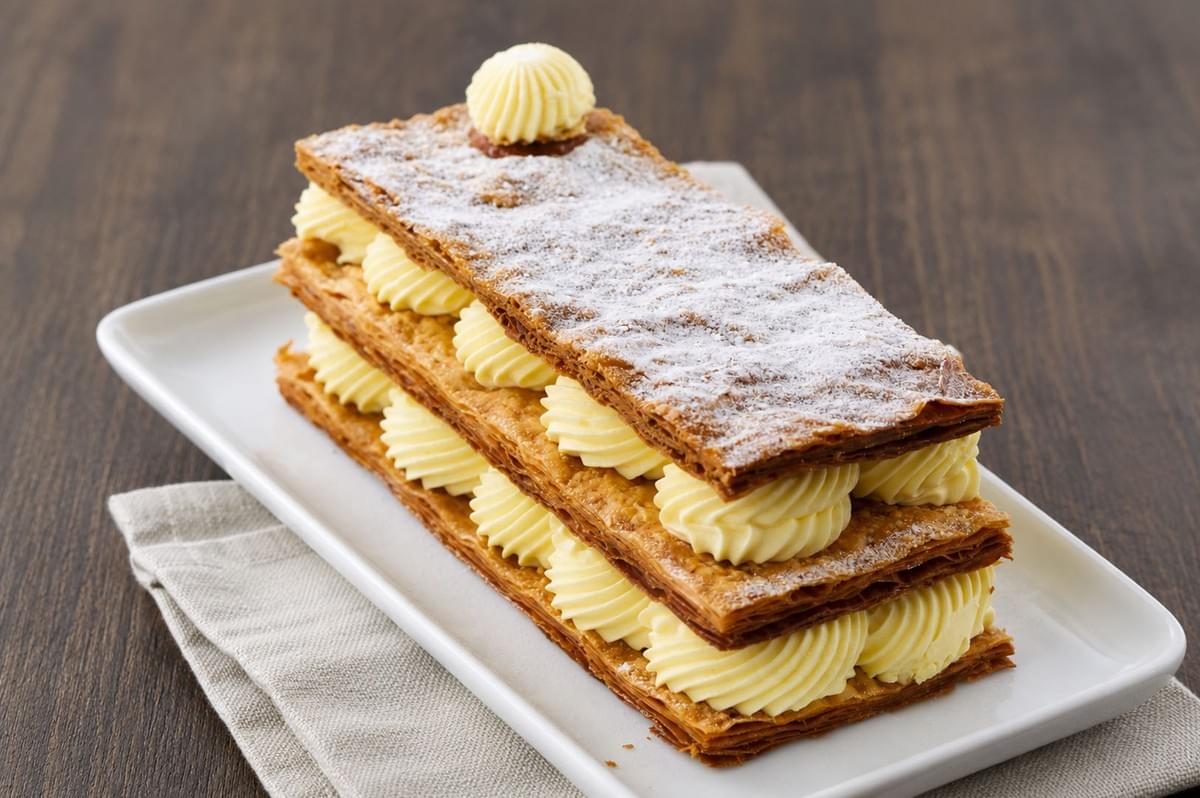 Classic Mille-Feuille with three layers of flaky puff pastry and piped vanilla cream, dusted with powdered sugar on a white rectangular plate Classic Mille-Feuille with three layers of flaky puff pastry and piped vanilla cream, dusted with powdered sugar on a white rectangular plate