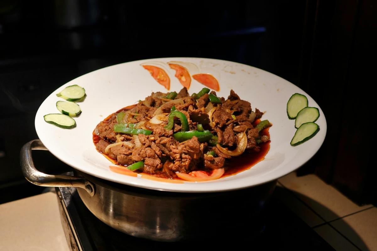 A large white bowl resting on a metal stand contains a savory stir-fry of sliced meat, onions, and green peppers coated in a rich dark sauce. Slices of fresh cucumber and tomato are arranged decoratively along the wide rim of the dish, framing the warm meal. A large white bowl resting on a metal stand contains a savory stir-fry of sliced meat, onions, and green peppers coated in a rich dark sauce. Slices of fresh cucumber and tomato are arranged decoratively along the wide rim of the dish, framing the warm meal.