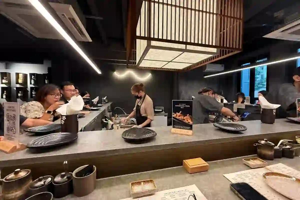 The image shows the bright storefront of a Putien restaurant, featuring a large illuminated sign and warm wood paneling. Inside, several patrons are seated at tables or waiting near the entrance of the modern, well-lit dining area. The image shows the bright storefront of a Putien restaurant, featuring a large illuminated sign and warm wood paneling. Inside, several patrons are seated at tables or waiting near the entrance of the modern, well-lit dining area.