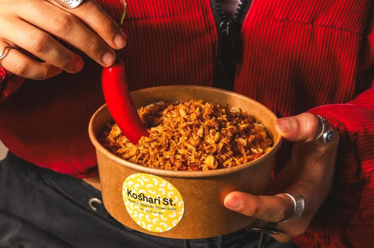 Takeaway bowl of Egyptian koshari from Koshari St., topped with crispy fried onions and held with a red chili pepper. Takeaway bowl of Egyptian koshari from Koshari St., topped with crispy fried onions and held with a red chili pepper.