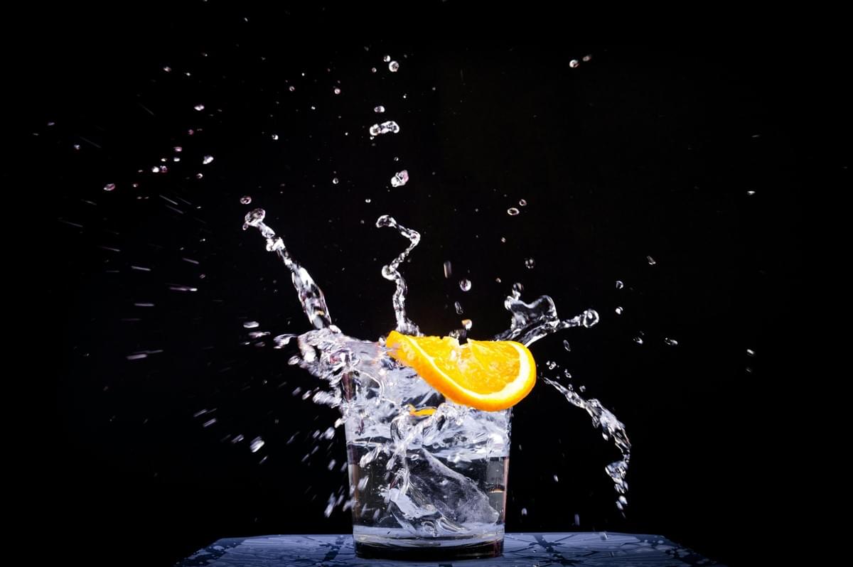An orange slice drops into a glass filled with water and ice cubes, creating a dramatic, dynamic splash. In the image, individual water droplets and streams are captured mid-air against a solid black background. An orange slice drops into a glass filled with water and ice cubes, creating a dramatic, dynamic splash. In the image, individual water droplets and streams are captured mid-air against a solid black background.