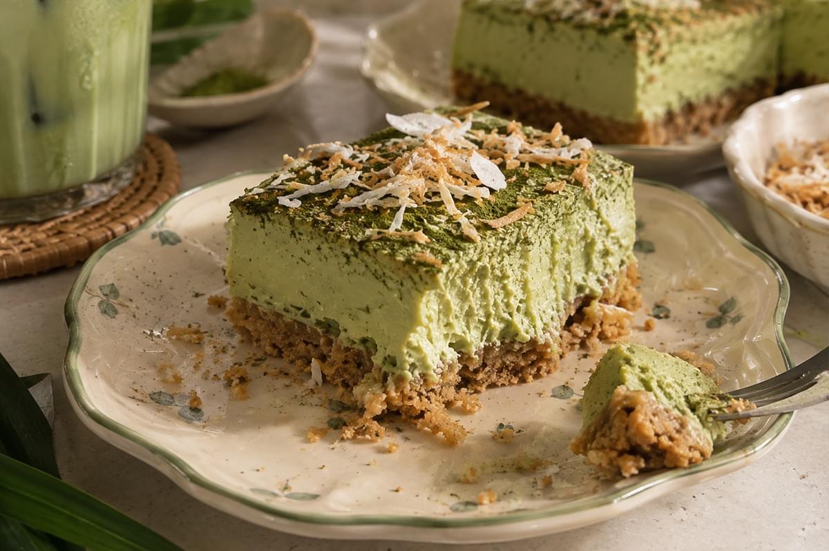 A close-up shot focuses on a single slice of pandan matcha cheesecake bar served on a vintage-style scalloped plate, showing its thick graham cracker crust and smooth, creamy pale-green filling topped with fine matcha powder and toasted coconut flakes. A fork picks up a small piece from the edge, while a tall glass of iced matcha latte and fresh pandan leaves sit softly blurred in the background. A close-up shot focuses on a single slice of pandan matcha cheesecake bar served on a vintage-style scalloped plate, showing its thick graham cracker crust and smooth, creamy pale-green filling topped with fine matcha powder and toasted coconut flakes. A fork picks up a small piece from the edge, while a tall glass of iced matcha latte and fresh pandan leaves sit softly blurred in the background.