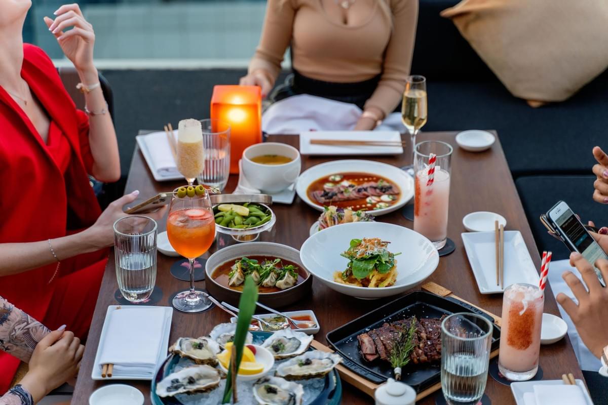 In the image, a group of friends is gathered around an outdoor dining table lavishly spread with gourmet dishes like fresh oysters, pan-seared steak, and dumplings. Brightly colored cocktails, elegant glassware, and smartphones fill the space, capturing a lively, upscale social dining experience. In the image, a group of friends is gathered around an outdoor dining table lavishly spread with gourmet dishes like fresh oysters, pan-seared steak, and dumplings. Brightly colored cocktails, elegant glassware, and smartphones fill the space, capturing a lively, upscale social dining experience.