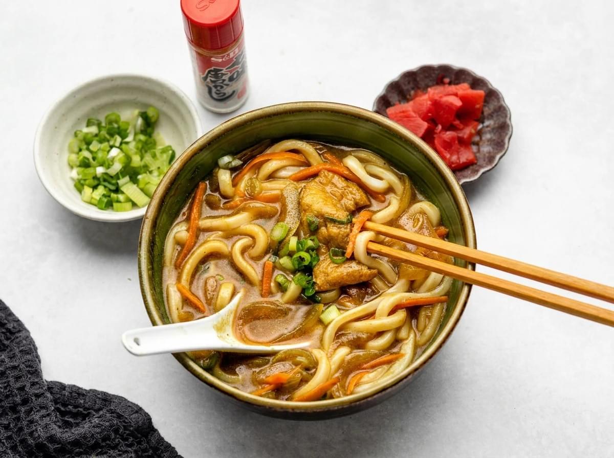Top-down view of curry udon with chopsticks, accompanied by green onions and pickled ginger sides. Top-down view of curry udon with chopsticks, accompanied by green onions and pickled ginger sides.