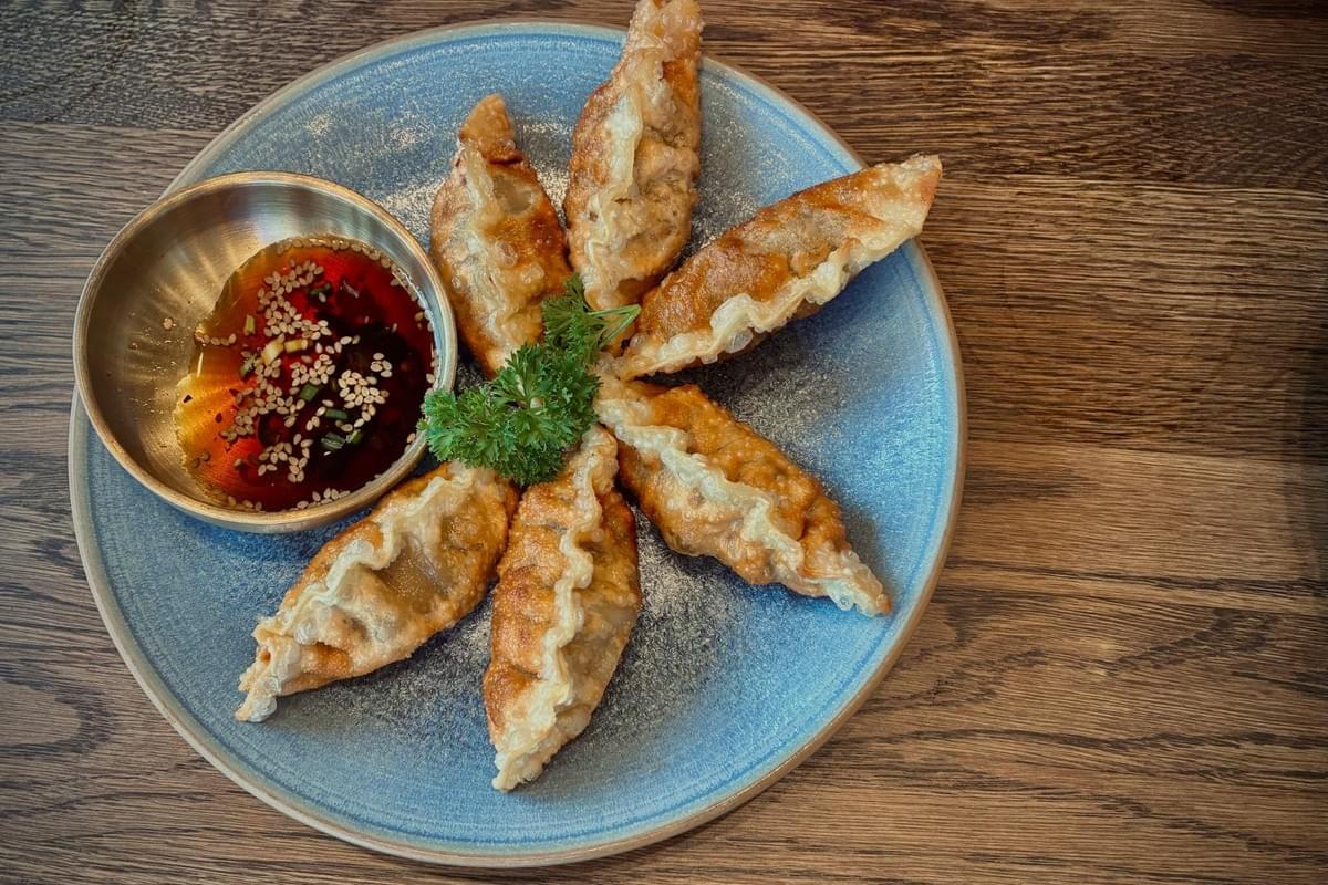 A top-down shot shows six crispy, golden-brown fried dumplings arranged in a star pattern on a textured blue plate next to a small metal bowl of dipping sauce topped with sesame seeds. The plate rests on a dark wooden table, with a small sprig of fresh parsley placing a touch of green at the center of the dish. A top-down shot shows six crispy, golden-brown fried dumplings arranged in a star pattern on a textured blue plate next to a small metal bowl of dipping sauce topped with sesame seeds. The plate rests on a dark wooden table, with a small sprig of fresh parsley placing a touch of green at the center of the dish.